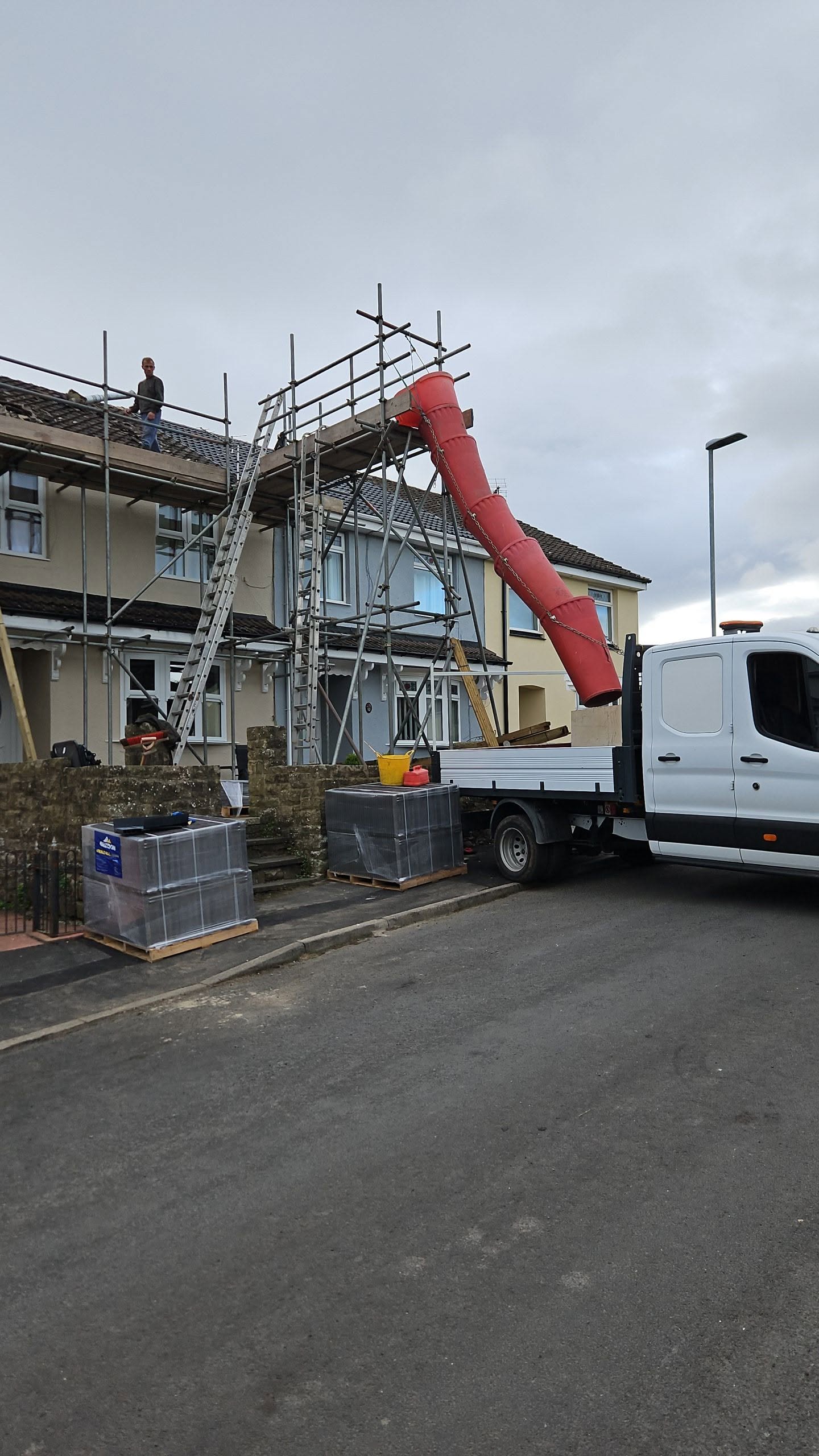 A flatbed truck and a red waste chute beside a scaffolded terrace